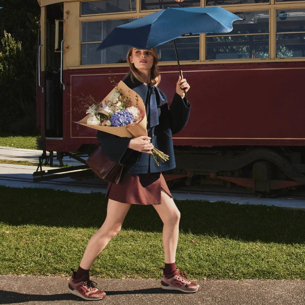 A woman walking outdoors on a sunny day, holding a blue umbrella and a bouquet of flowers, with a vintage red and cream tram in the background.